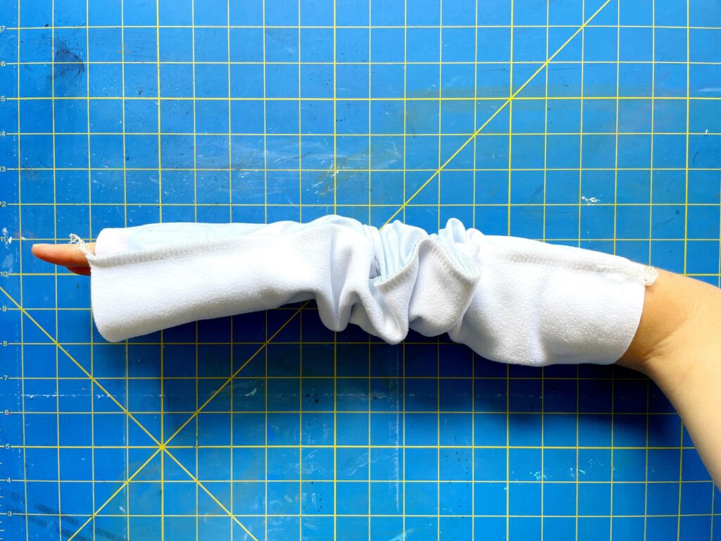 A hand is in between two pieces of fabric that are sewn together. The tips of the fingers are seen at one end, while the wrist is seen at the other end. The fabric is scrunched up in the middle. A blue and yellow cutting mat is the background.