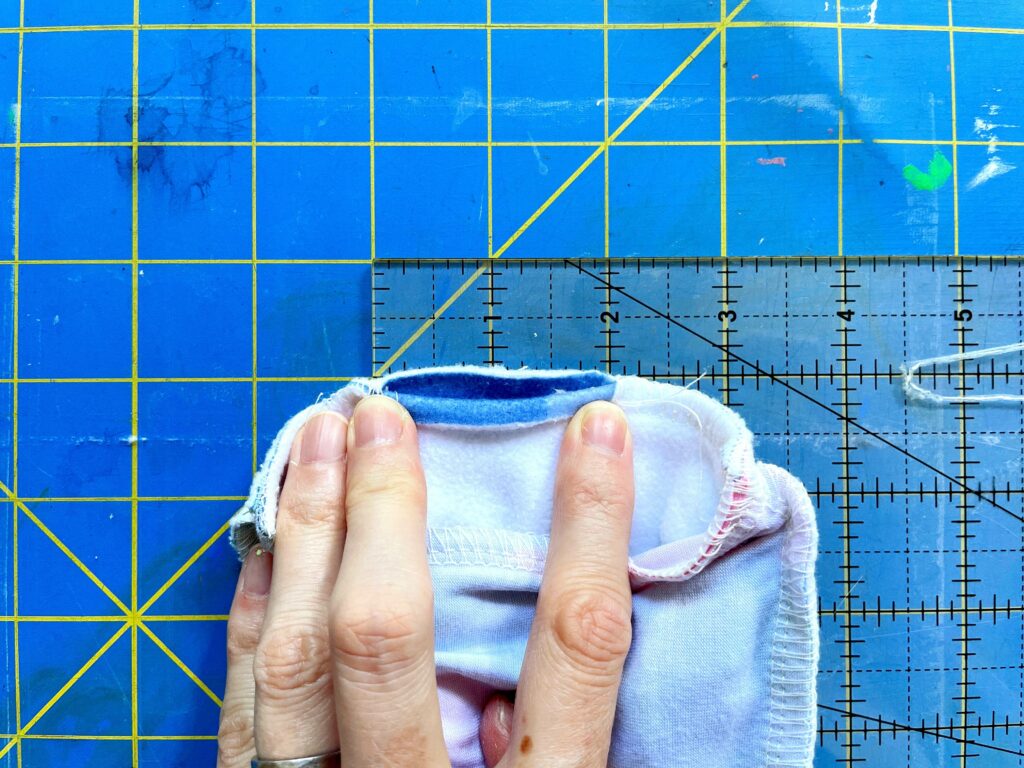 A hand holding down an opening of two sewn pieces of fabric. Two fingers in particular rest at the edge of the opening to indicate its width at two inches by using a clear measuring board. A blue and yellow cutting mat is the background.