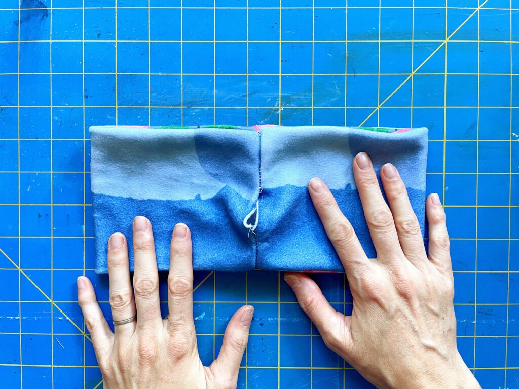 Two hands rest on a piece of blue folded fabric. The stitch where the two ends of fabric have been sewn together is in the space between the hands. The opening between the top and bottom of the stitch is in the middle. A blue and yellow cutting mat is the background.