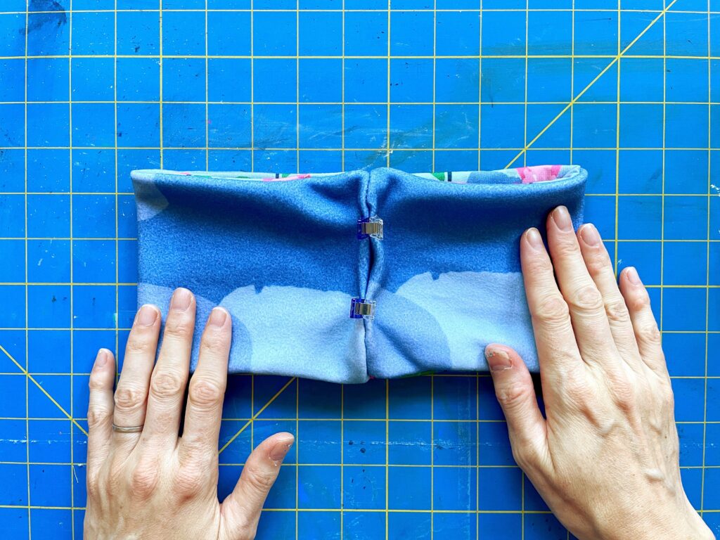 Two hands rest on the ends of the fleece fabric headband. Clips are in the center where the shorter sides were sewn together. A blue and yellow cutting mat is the background.