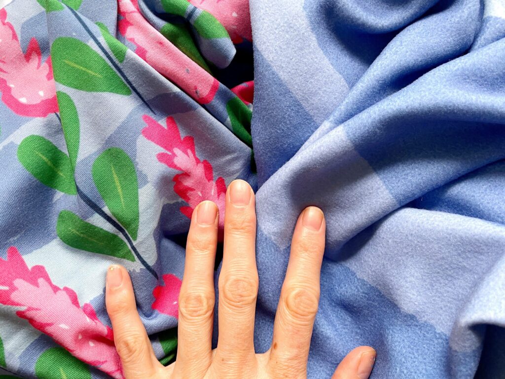 Two fabrics lay lightly piled under a hand. The left fabric has pink flowers with green leaves. The right fabric is made of two different tones of blue.