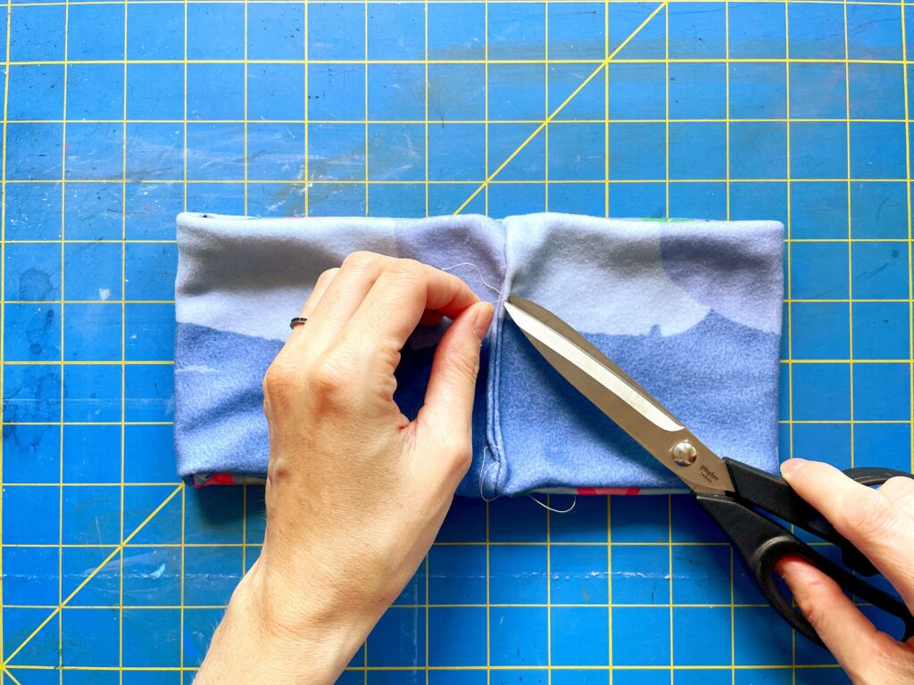 One hand is holding a piece of excess thread on the blue fleece headband while the other hand is clipping it with black fabric scissors. A blue and yellow cutting board is the background.