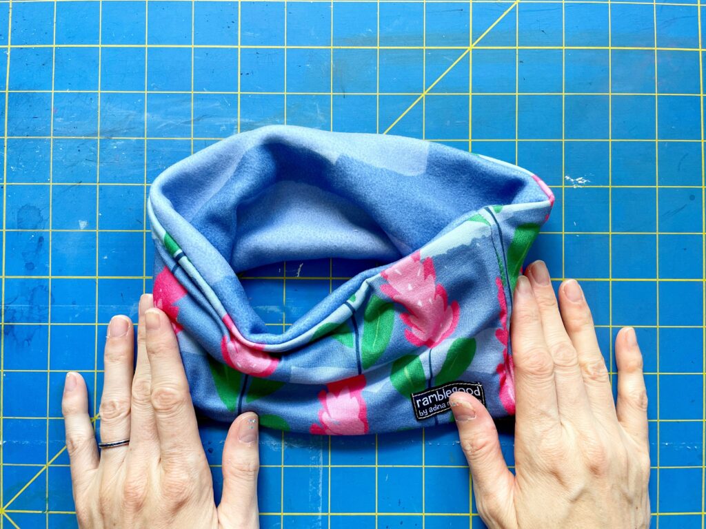Two hands rest at the edge of the finished headband. The pink and green flowered fabric is exposed from the outside, as the blue polartec fleece is visible from the inside. A blue and yellow cutting mat are the background.
