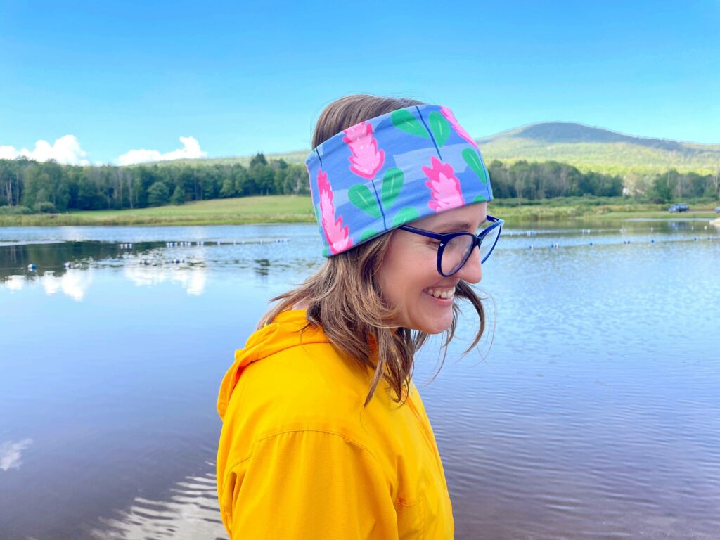 A woman wearing a blue headband with pink flowers is turned to the right. She’s wearing dark blue glasses and a yellow hoodie. A lake, trees and a mountain are behind her.