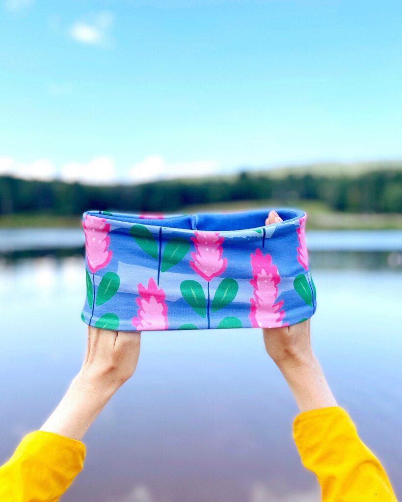 Two hands are inside the ends of a fleece headband to show what it looks like. The bottom of the hands through a quarter of the forearms is visible, followed by yellow sleeves. The outside of the headband is blue with pink flowers and green leaves. The inside is blue. We see an out-of-focus lake and skyline as the background.