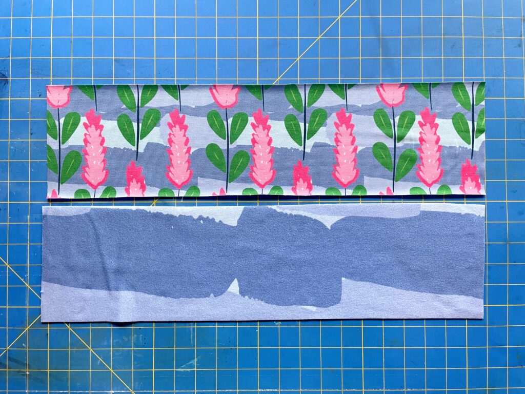 A blue striped fabric with pink flowers and green leaves lays above a fabric with different streaks of blue in different hues on a blue and yellow cutting mat.