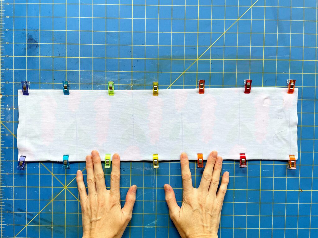 Two hands hold down fabric that is turned right side in and secured with clips. A blue and yellow cutting mat makes up the background.