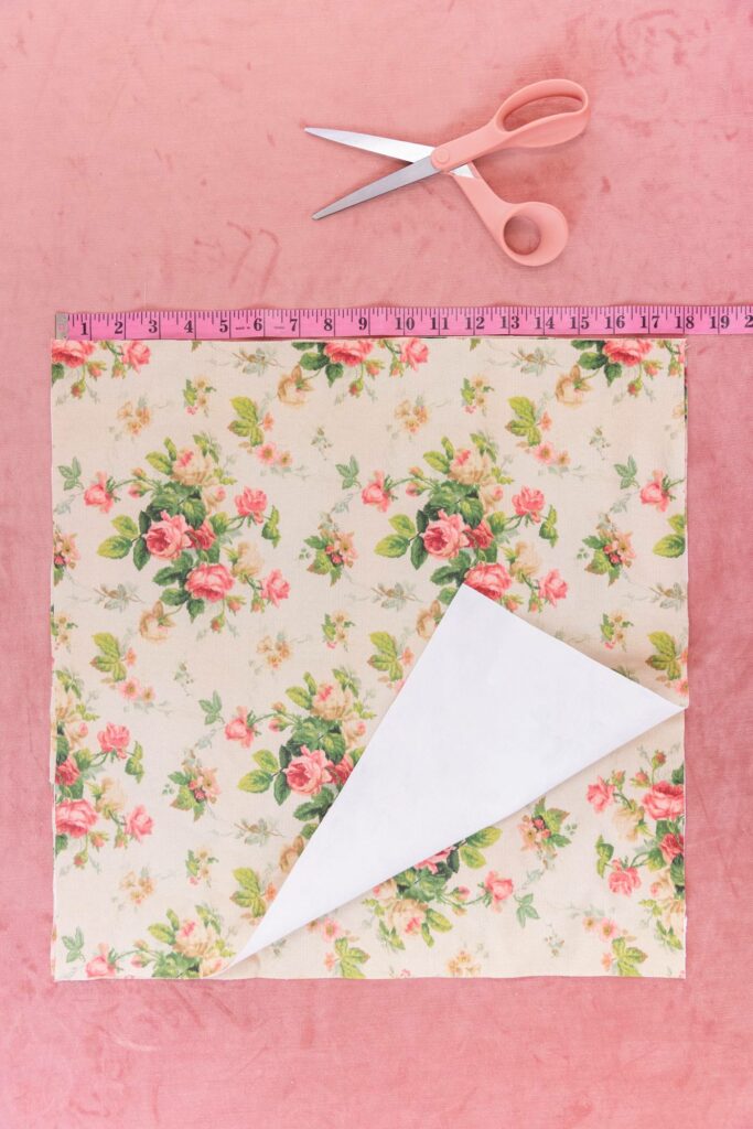 Two pieces of floral fabric rests beside a pink measuring tape and peach-colored pair of scissors. Pink carpet surround the materials in the background.