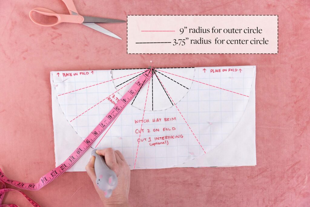 A circle template on grid paper is pinned down on top of a piece of folded fabric, with the design side in. A pink measuring tape is pinned at the top center of the folded fabric, extending out, to indicate how to measure the hat’s brim. Black and red dashes indicate where to measure the radius. Using a mouse-shaped pen, Keiko indicates where to draw lines to trace the template. A box to the top right of the photo indicates the red dashes are for measuring the outer circle and the black dashes are for measuring the inner circle.