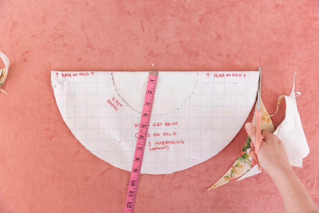 Following along the semi-circle template, Keiko cuts the fabric. A pink measuring tape pinned to the fabric extends from the top-middle of the semi circle.