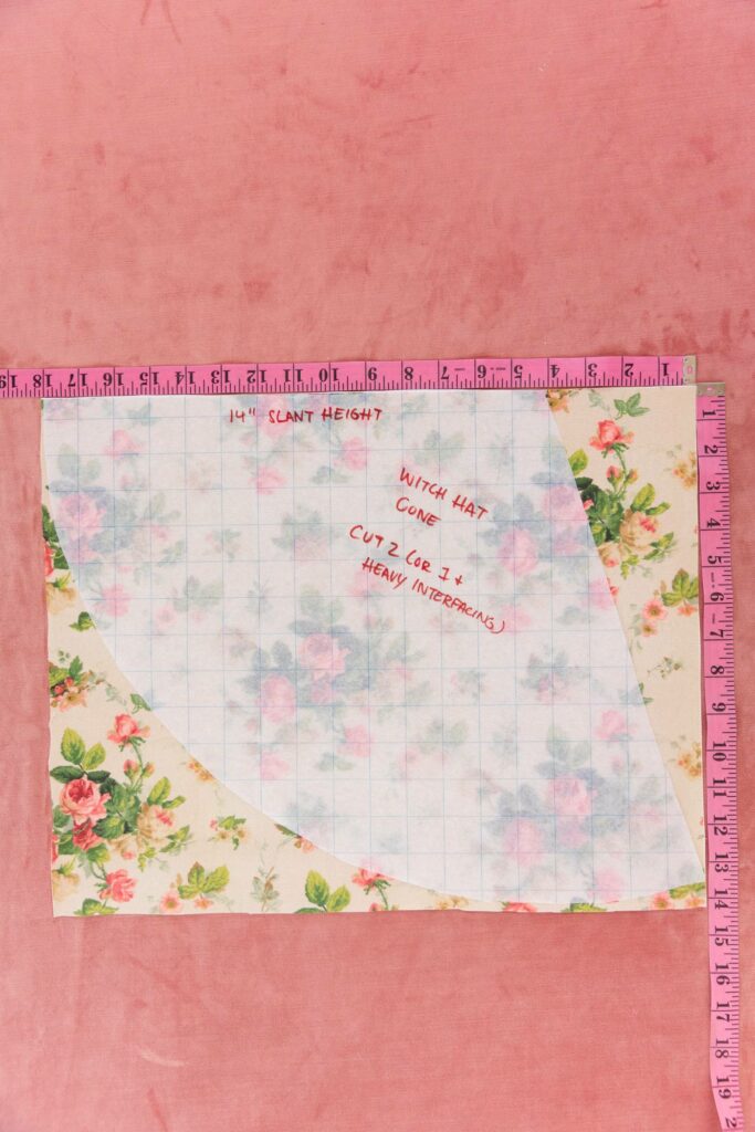 Translucent grid paper lays on top of a cream floral piece of fabric with pink roses and green leaves and stems. Two pink measuring tapes are beside the fabric measuring its length and width. A pink carpet is behind as the background.