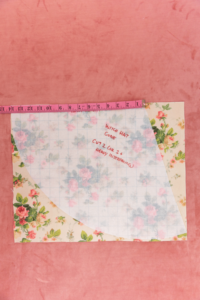 A translucent grid paper is on top of a cream floral fabric with pink roses and green leaves. One pink measuring tap lays on the edge of the fabric measuring out 14-inches. The background is a pink carpet. The measuring tape moves across the grid paper until it reaches the edge.