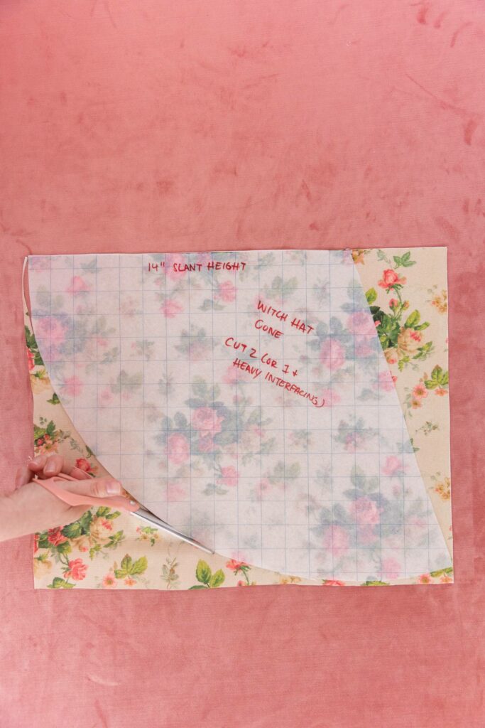 Keiko cuts a cream fabric with pink roses and green leaves in the cone shape of the translucent grid paper resting on top of the fabric, with a peach-colored pair of scissors. A pink carpet is in the background.