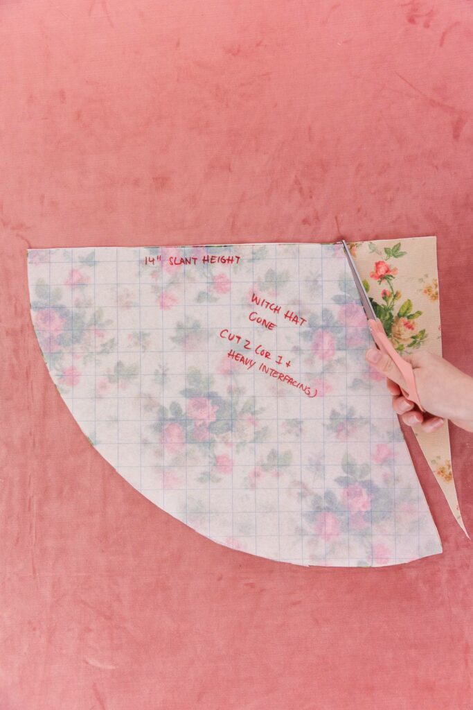 A hand cutting the remainder of crème floral fabric with pink roses and green leaves into the cone shape of the translucent grid paper. Pink carpet is used as the background.