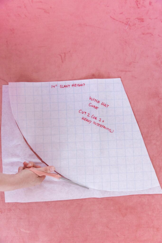A white piece of interfacing is underneath a translucent grid paper template. Keiko is cutting the interfacing in the shape of the grid paper template with a peach-colored pair of scissors. Pink carpet is used in the background.