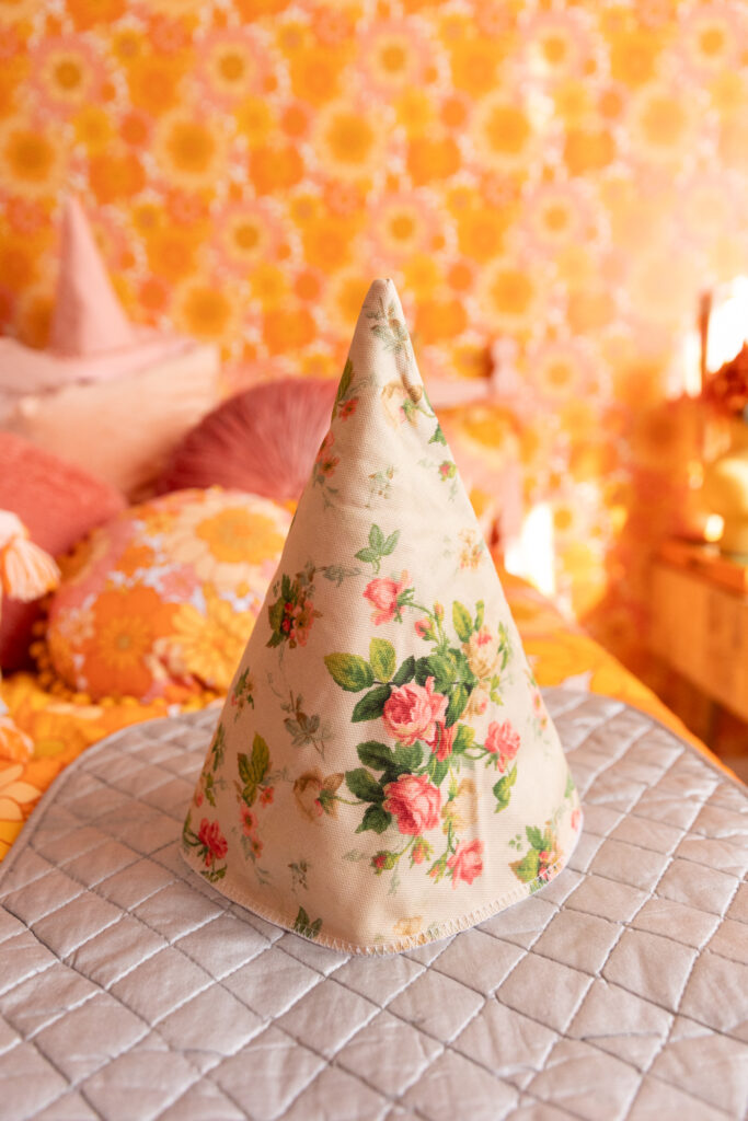 A cream colored cone with pink roses and green leaves sits on a silver ironing mat. The mat is resting on a bed with retro floral bedding. Out of focus, to the left are throw pillows in different shades of pink. Wallpaper, that matches the bedding is on the wall behind everything.