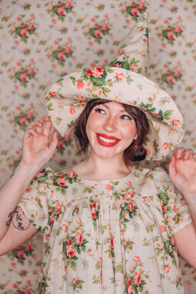 Keiko wearing a floral witch hat in a matching dress in front of a backdrop of the same design. She's smiling with the tips of her fingers touching the brim of her hat. 