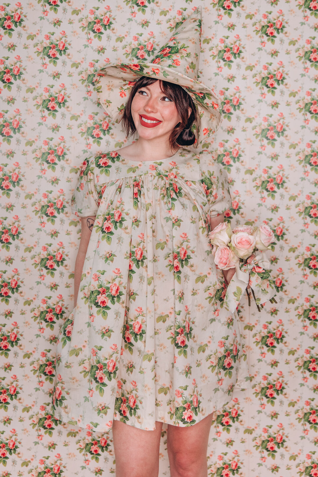 Keiko in a floral witch hat with a matching dress in front of a backdrop made of the same design. She's holding flowers in her right hand, looking up to the left.