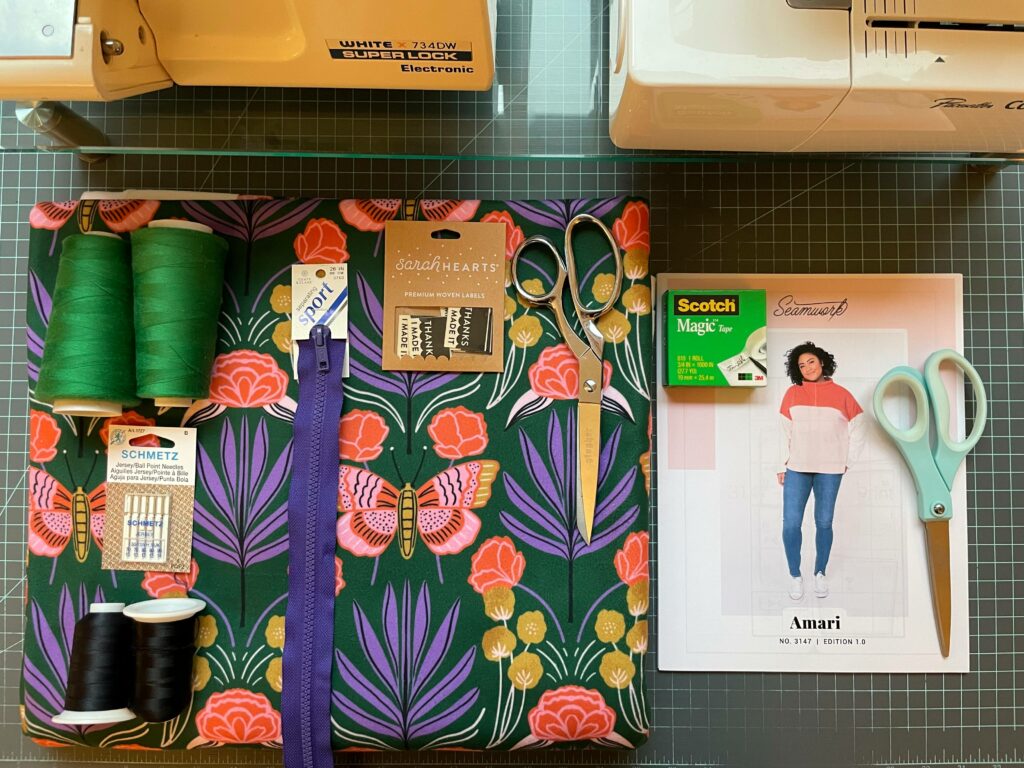 A green grid surface lays underneath supplies to make a fleece pullover. Green thread, black thread, sewing needles, a sports zipper, clothing labels and fabric scissors lay on top of a green botanical fabric. To the right side of that are another pair of scissors, a sewing template and tape. A sewing machine and serger are in the top left and right corners. 