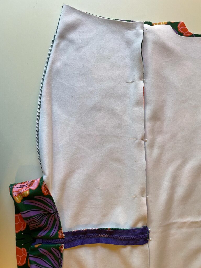 The inner neckband has been stitched to the rest of the jacket which is turned design side in. The zipped is closed and sewing pins connect the yoke to the front fabric that needs to be stitched. A white surface is in the background.