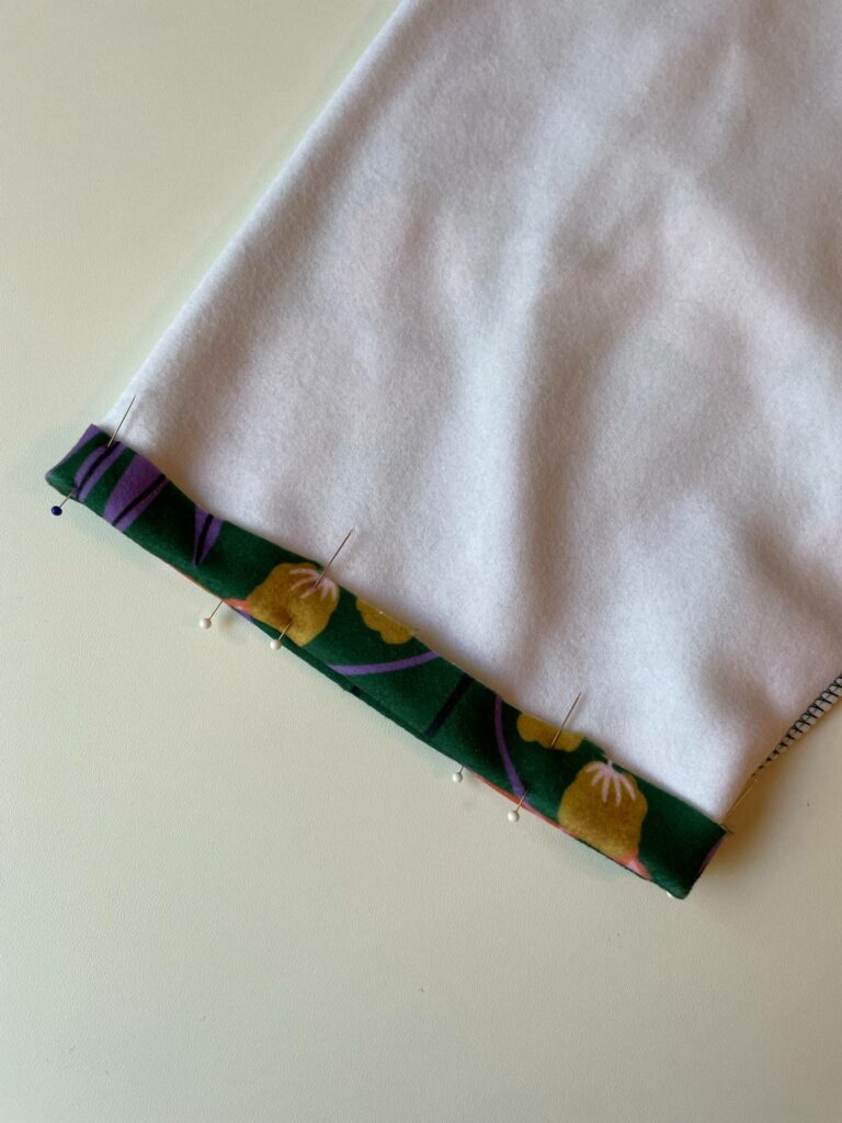A closeup on the end of the sleeve shows it’s folded back with sewing pins placed along the edge in preparation to hem the sleeve. The folded back edge shows the design. A white surface is in the background.