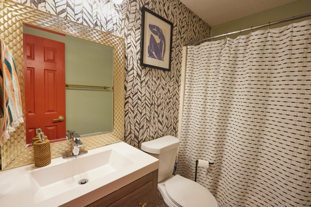 A bathroom with a gold, textured mirror sits in front of black and white leaf designed wallpaper. A framed picture is hung above the toilet. A black and white shower curtain covers the shower. A red door and sage green wall are in the reflection of the mirror. A hand towel hangs from the hook on the left wall beside the bathroom sink and mirror.