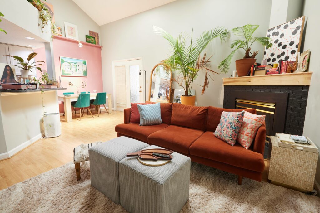 Two grey ottoman sit in front of a rust orange couch with two throw pillows on each corner. A cream and light brown rug sits underneath. Two potted plants, and two mirrors sit behind to the left. A black fireplace with stacked books and vases are to the right. The dining room area is visible to the far left. A pink accent wall with a painting faces a light wooden table with green chairs. A bar area leading to the kitchen supports plants and a record player. 
