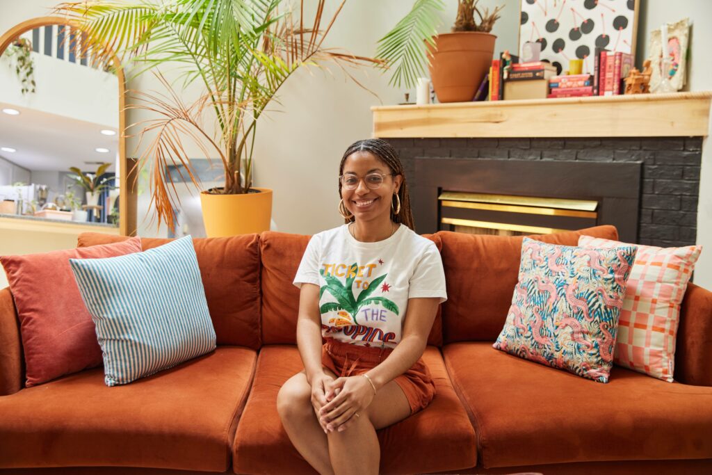 Kristina sitting in the middle of her rust brown couch in a white t-shirt and rust colored shorts. Four throw pillows of different designs sit between her. A potted plant sits behind her. In Leaning on the wall is a oval full body mirror beside a smaller rectangular mirror. In the other corner is a black brick fireplace with books, candles and a canvas print resting on the mantle. 