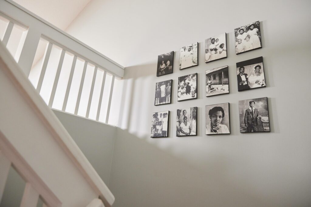 Vintage family photos in black and white are on twelve square canvas on a light grey wall. A handrail and beam are to the left of the wall indicating the staircase location.