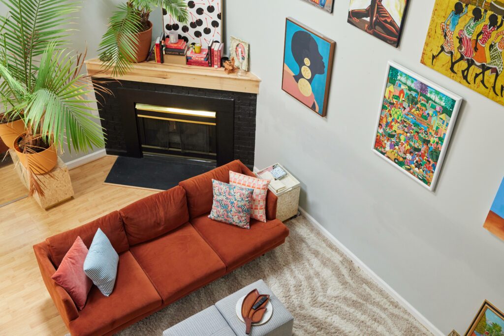 From an angled aerial view, we see two throw pillows each on the corners of a rust orange couch. A cream and tan colored rug are at the foot of the couch underneath two square ottomans. A black fire place with candles, a plant and stacked book on the mantle are behind the couch. Colorful artwork hangs from the right wall. 