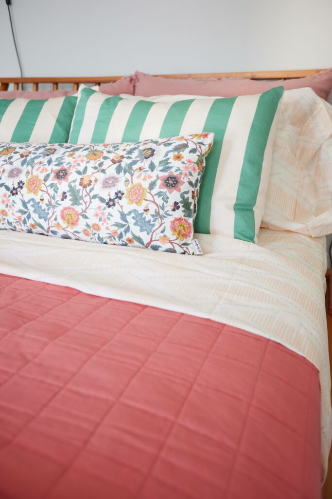 A corner close up of a bed showing the detail in blush red colored comforter, flowered lumbar pillow, cream and green striped pillows and orange and tan sheet set with matching pillow shams. A wooden headboard sits behind the pillows, followed by a white wall.