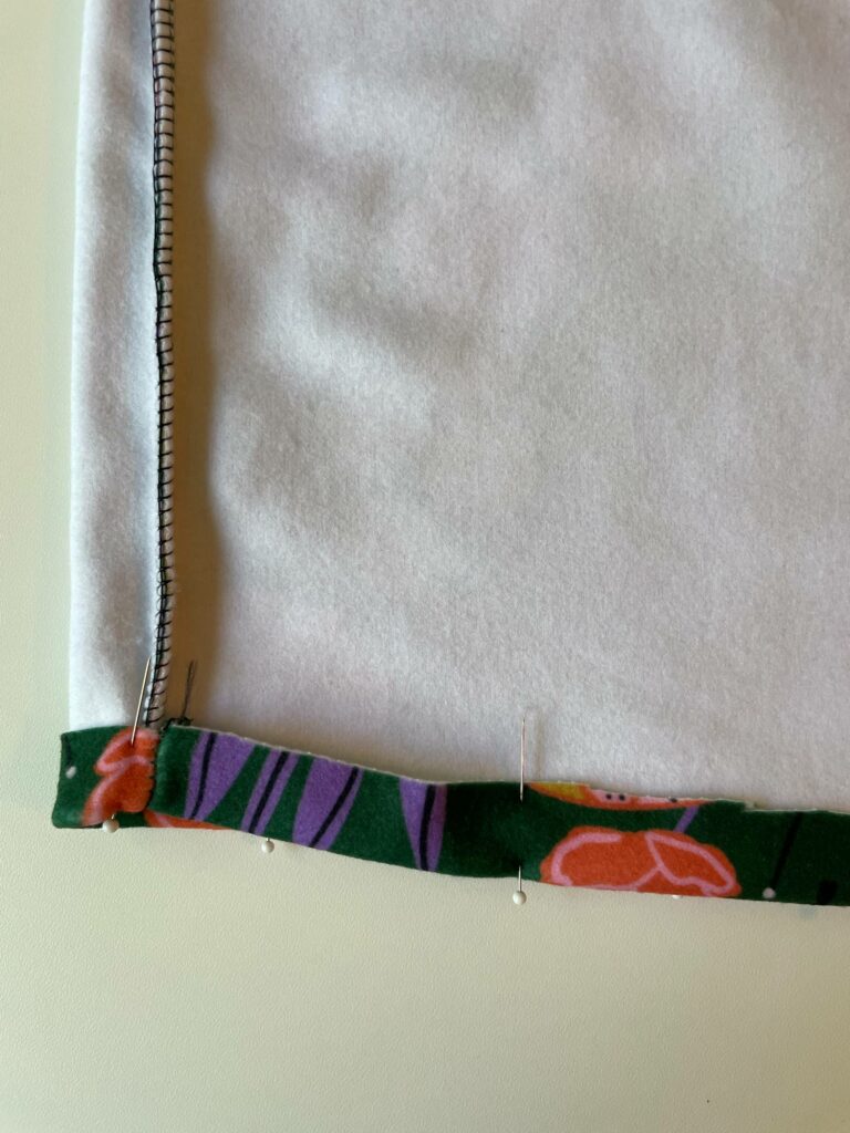 A close-up of a downward-pointed sleeve. It is turned inside out where the sleeve stitches are visible. Sewing needles are placed where the end of the sleeve will be hemmed. The sleeve is on a white surface that is in the background.