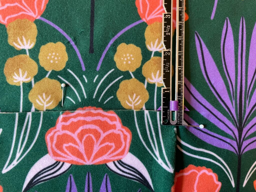 A ruler and two sewing pins are on a botanical piece of fabric. There's a break in the fabric indicating on piece is on top of another. 