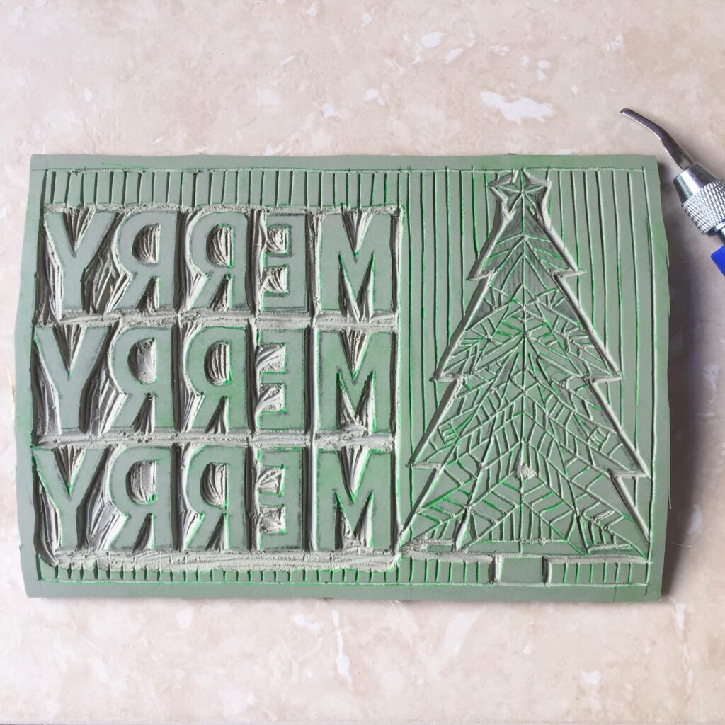 A green block print of the words "Merry" stacked in three rows beside a christmas tree. A concrete surface is in the background.