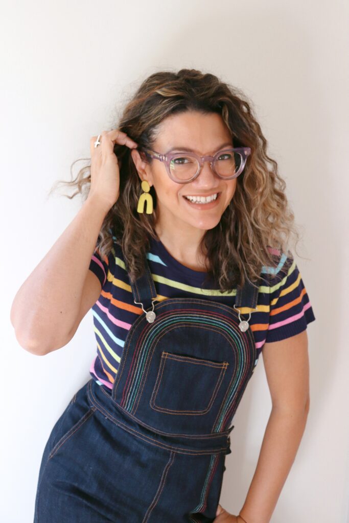 Amarailys in dark blue denim overalls with a striped shirt. She's wearing yellow earrings and purple glasses. She's smiling and pushing hair behind her eat with her right hand.