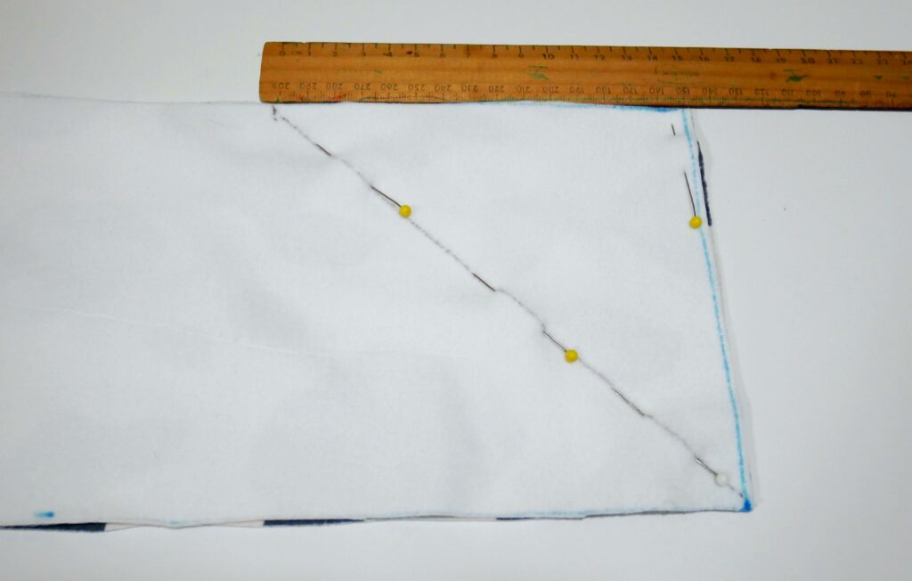 The wrong side of the fabric is turned up and a diagonal line has been drawn from the bottom right corner up 5.9” or 15 cm. Pins are placed along the diagonal line. A brown wooden ruler is at the top of the fabric, making sure the line ends at 15 cm.