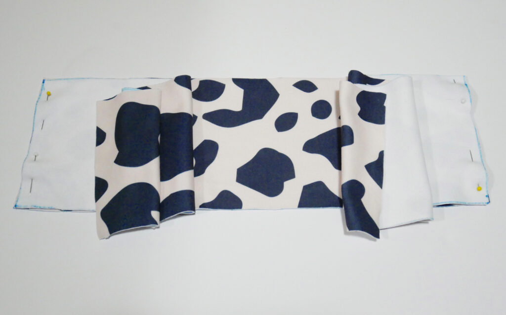 The Inner Bolster pieces have been cut out of fabric that is white with black cow-print shapes. The two center pieces have been pinned together at the edges wrong side out. The longer side pieces are laying on top of the shorter center piece.