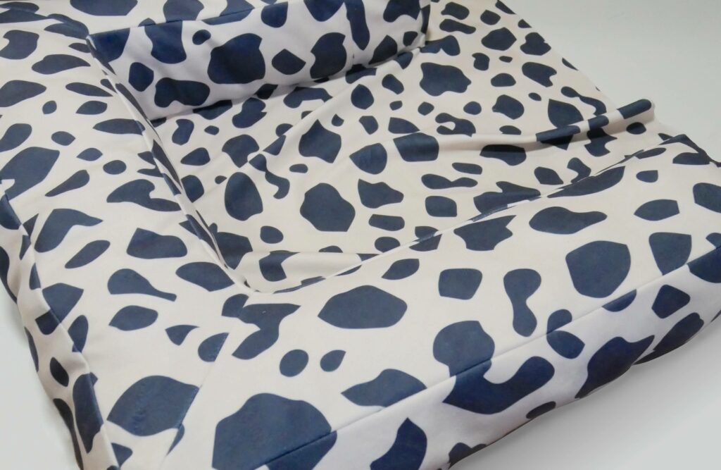 The bolsters are now sewn to the base of the dog bed and stuffed with fabric. The base of the bed still needs to be filled with fabric. The fabric is white with black cow-print shapes.