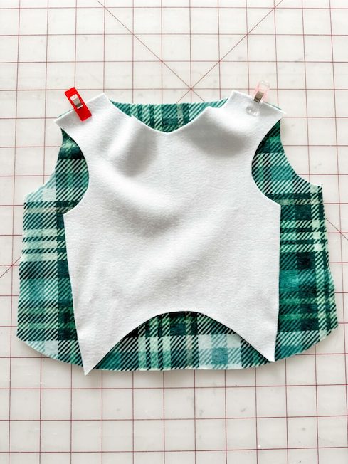 The wrong side of a green-and-white plaid fabric piece lays on top of another green-and-white fabric piece that is design side up. They are held together using sewing clips. The fabric lays on a white cutting mat with a red grid.