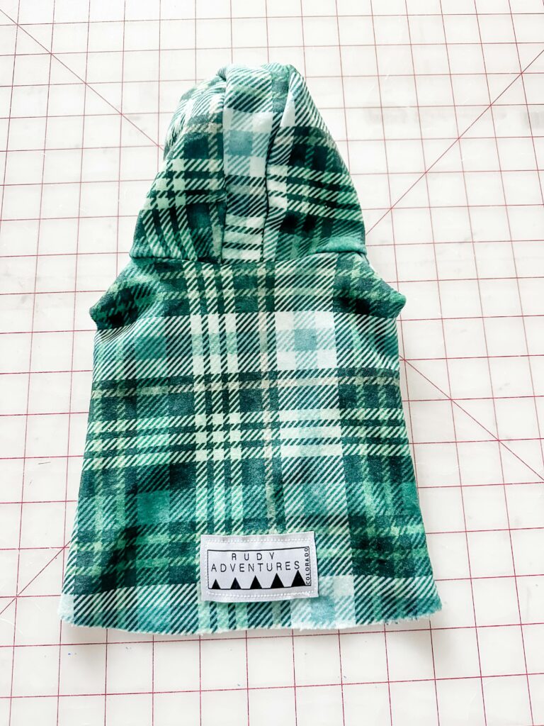 A plaid green-and-white dog coat is on a white cutting mat with a red grid with the back of the hood facing up. The inside of the hood is white with red dots and evergreen leaves. A small “Rudy Adventures” label is in the bottom center.