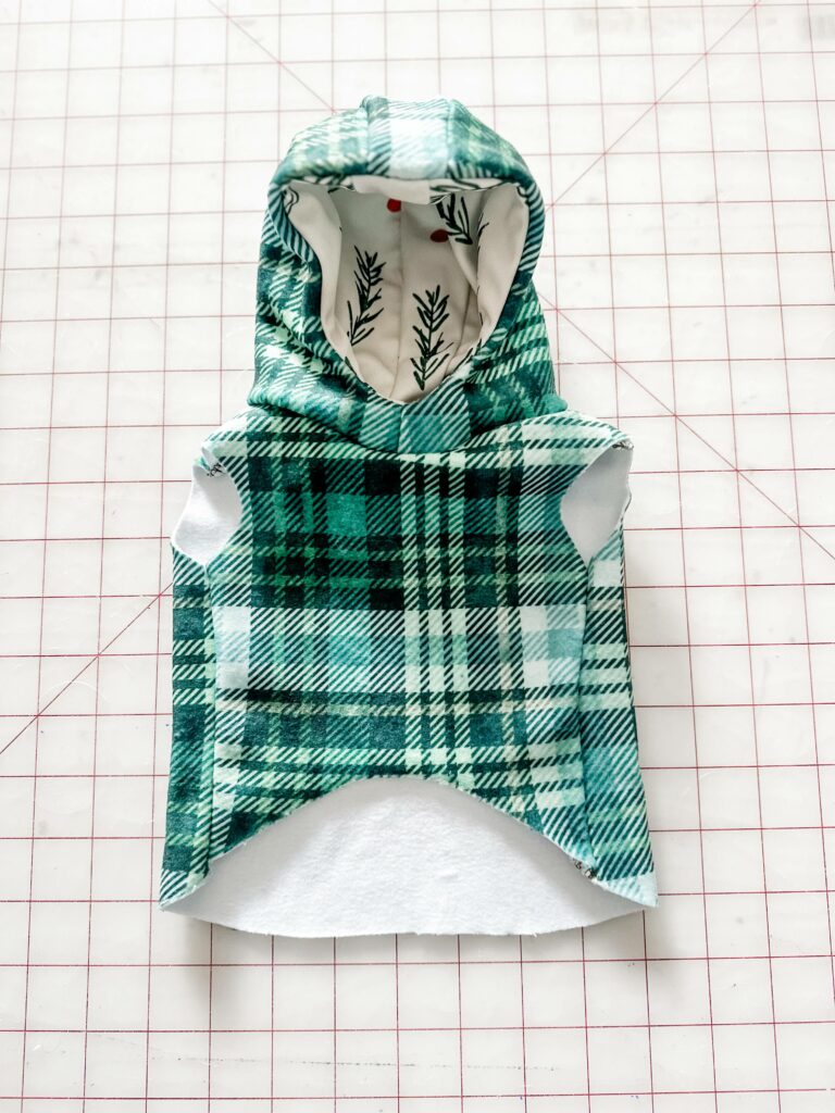 A plaid green-and-white dog jacket is on a white cutting mat with a red grid with the front of the hood facing up. The inside of the hood is white with red dots and evergreen leaves.