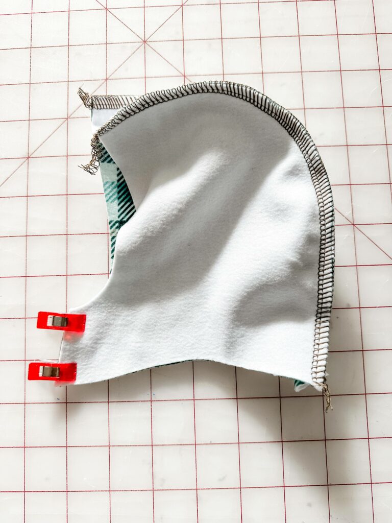 One of the hoodie’s side pieces is shown design side down and sewn to the curve of the center piece. Two sewing clips are at the hoodie’s small front center piece holding the two side pieces together. The fabric lays on a white cutting mat with a red grid.