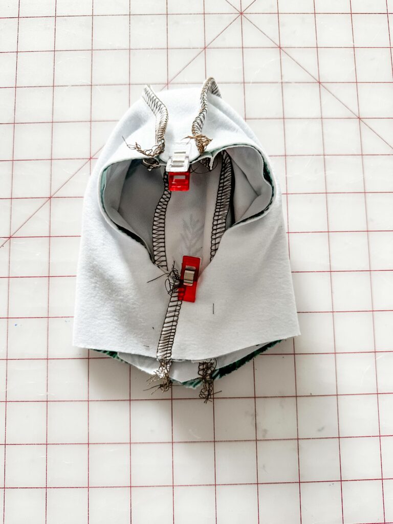 The exterior hoodie piece is turned design side in while the interior hoodie piece is turned design side out. The exterior hoodie piece is placed inside the interior hoodie piece. Sewing clips secure the two pieces at the center top and bottom where the dog's head would go. The fabric lays on a white cutting mat with a red grid.