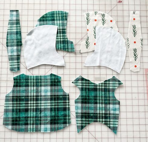 Plaid green hoodie and bodice fabric pieces lay to the lefthand side on a white cutting mat with a red grid. Fabric pieces for the hoodie in a design with a white background and red dots and evergreen leaves lay to the righthand side of the cutting mat.