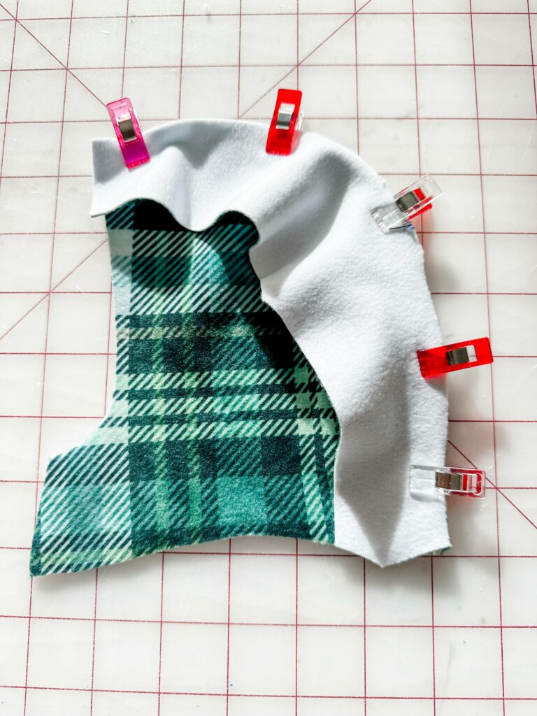 A side piece of the hood is design side up and is clipped to the center piece using sewing clips. The hood’s center piece is design side down. The fabric lays on a white cutting mat with a red grid. The design on the fabric is a green-and-white plaid.