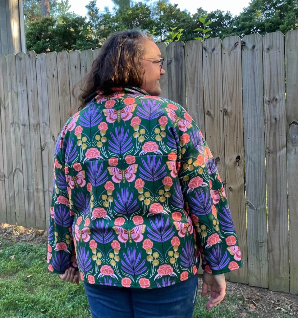 MaryAshlyn showing off the back of her finished Amari jacket. The consistency of the directional pattern is evident. A fence is in front of her followed by the edge of a house and some trees.