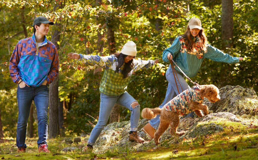 Three people are walking in the woods. The person at left is walking through the grass, the two people on the left are walking over large rocks along with a goldendoodle. The people are wearing jeans and quarter-button fleece pullovers in bright designs. The dog is wearing a fleece coat with a design featuring mountains ranges drawn in muted jewel tones