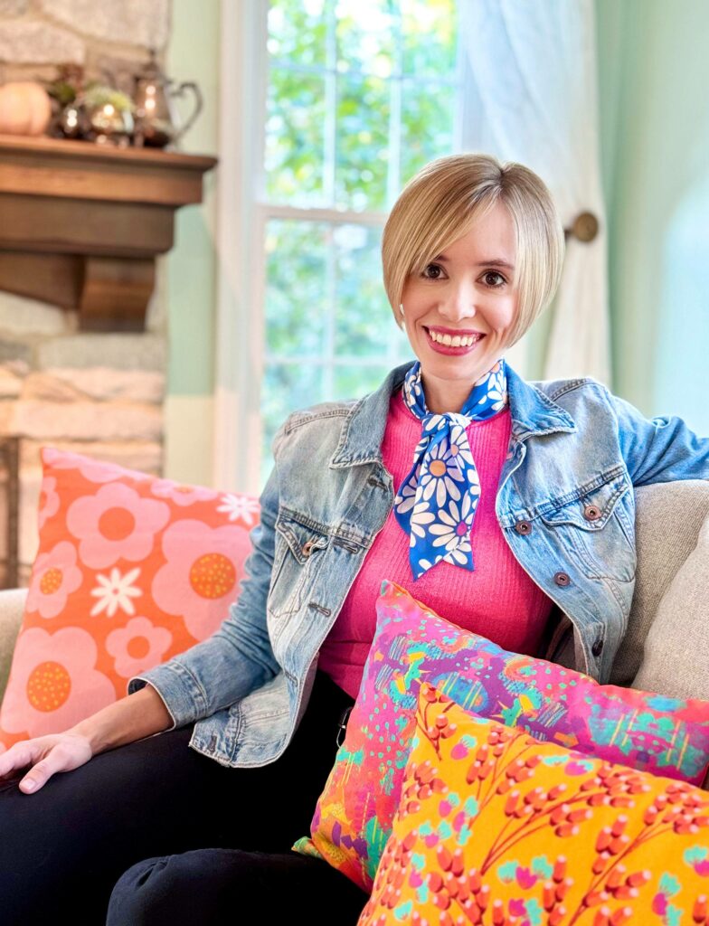 Katie Snow sitting on a couch surrounding by pillows in different designs. A fireplace is the back left corner. A window is to the right, with a curtain pushed to the corner. 