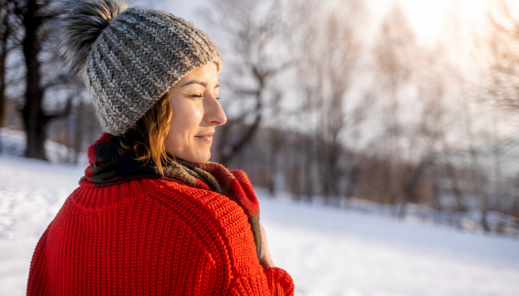 A woman stands outside in the winter with snow on the ground and bare trees behind her. She is bundled in a red sweater and brown hat, with eyes closed and a relaxed expression as the sun shines on her face.