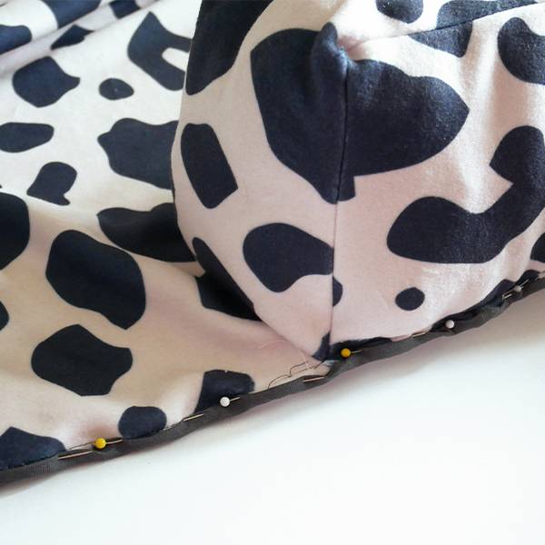 The rectangular bolster base has been flipped back over so that the fabric, which is white with black cow-print shapes is showing. This is a close up of the black bias tape being pinned around a bottom edge of the bolster base.